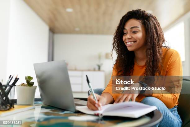 Schwarze Frau Sitzt Am Schreibtisch Und Verwendet Computerschrift Im Notizbuch Stockfoto und mehr Bilder von Lernen