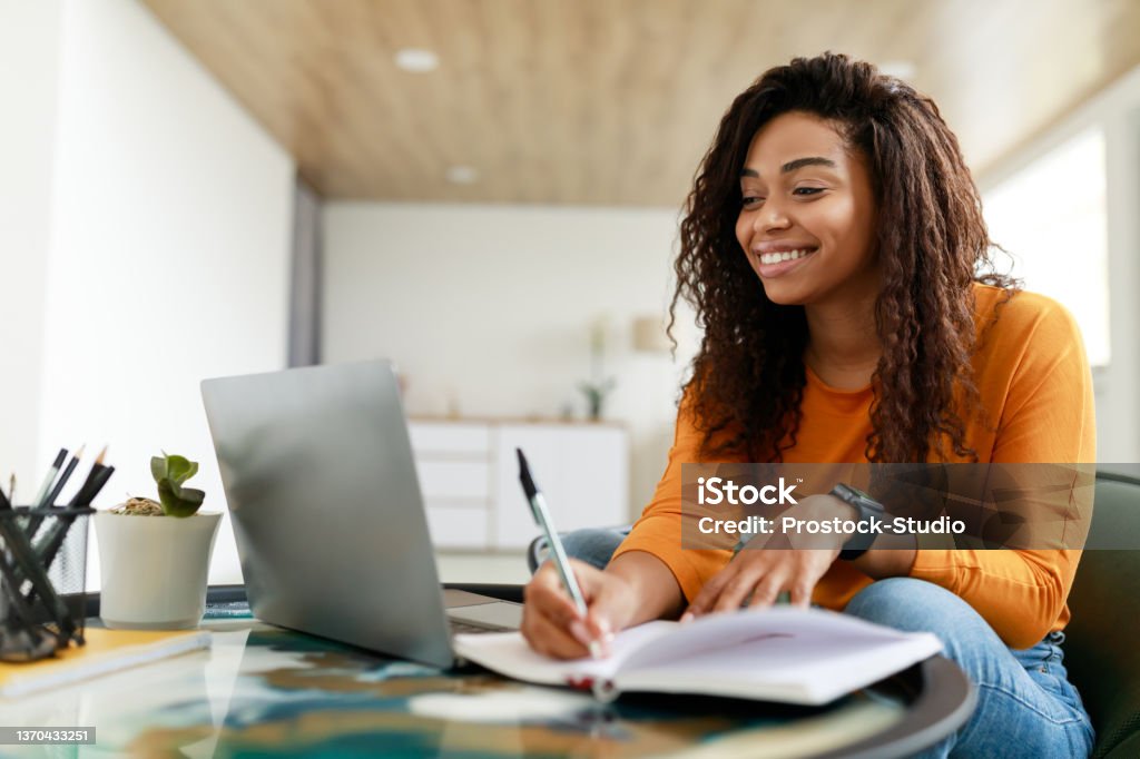 Black woman sitting at desk, using computer writing in notebook Note Taking Concept. Smiling young African American woman sitting at desk working on laptop and writing letter in paper notebook, holding pen and looking at screen. Happy millennial female using pc Studying Stock Photo