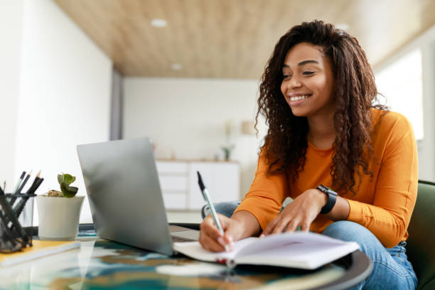 Black woman sitting at desk, using computer writing in notebook stock photo