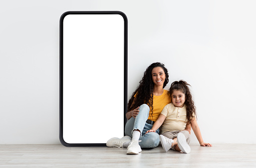 Happy arab mother and daughter sitting near big cellphone with empty white screen, advertising new cool app over light studio wall, mockup for your website design