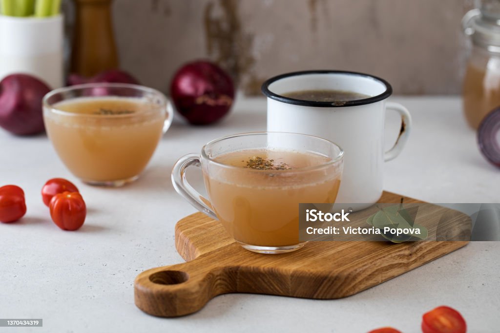 Homemade bone broth in mugs with spices on a wooden stick with bay leaf, tomatoes and onions. Homemade bone broth in mugs with spices on a wooden stick with bay leaf, tomatoes and onions. High quality photo Bone Broth Stock Photo