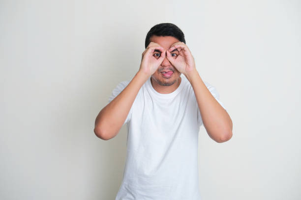 Adult Asian man making binoculars shape with his hands and looking through it with curious expression