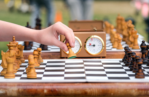 Close-up of a hand doing the opening move in a game of chess during a tournament with clock and another chess board blurred in the background, selective focus