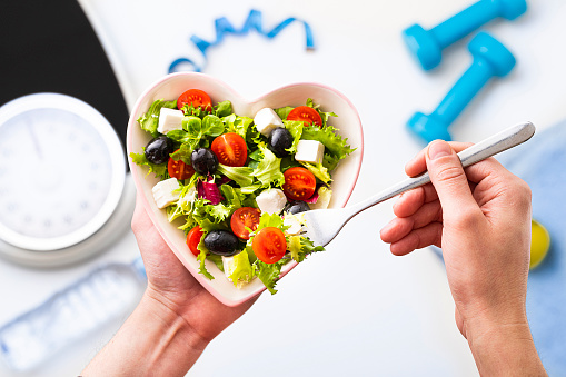 Top view of two human hands, one holding a heart shaped bowl full of salad and the other holding a fork. The main focus is on the bowl and at the defocused background are a bathroom scale, a tape mesure, two dumbbells, a water bottle and a towel.