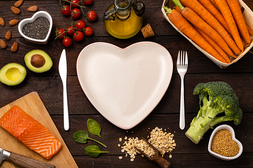 Top view of a heart shaped plate surrounded by a knife and a fork and some healthy food such as a salmon fillet, broccoli, oat flakes, chia and flax seeds, cherry tomatoes, carrots, almond, avocado, spinach and olive oil. The plate is empty so there is a useful copy space on it. All the objects are on a rustic wooden table.