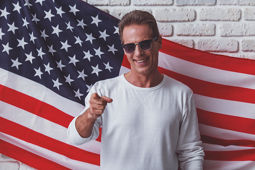 Handsome man in white sweatshirt and sunglasses is smiling, pointing and looking at camera while standing against American flag