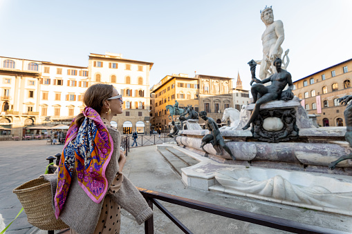 Young woman traveling famous italian landmarks in Florence city. Enjoying beautiful architecture and Neptuine fountain on Signoria square. Woman dressed in Italian style with colorful scarf in hair