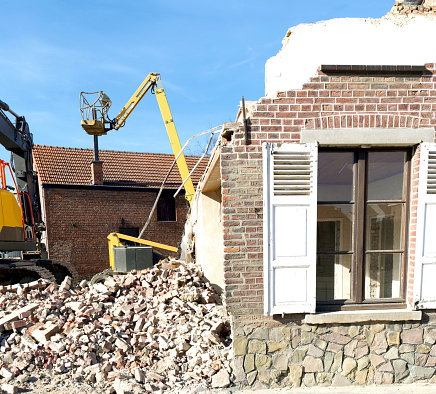 Dismantling a house more than 100 years old. Facade still remaining. Background hydraulic lift and excavator. Front heap of broken bricks laying on ground from demolition