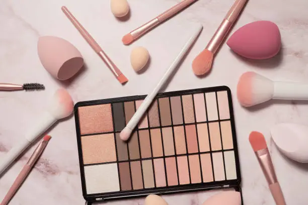 Photo of A nude eyeshadow palette and makeup artist's tools on a marble vanity. Brushes for powder, blush, eyebrows, shadows and sponges for concealer and foundation.