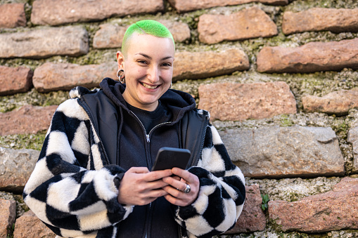 portrait of young authentic androgynous woman with short haircut and colored green, genuine smiling lesbian woman using smartphone, happy girl with piercing and street fashion dress code