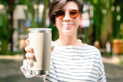 Happy, healthy and beautiful young Asian woman with sunglasses smiling, holding and showing single use coffee cup and reusable insulated tumbler with green tropical trees and leaves in background. Sustainable lifestyle and Zero waste concept.