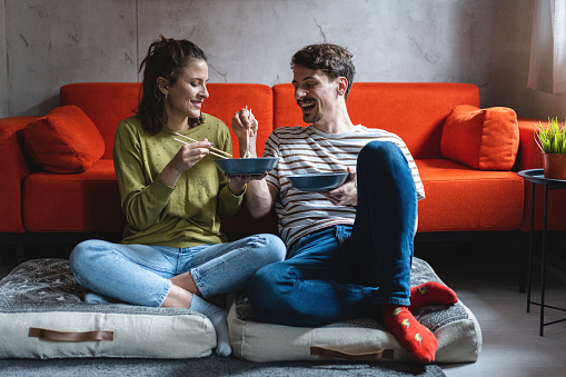 Cheerful young Caucasian couple enjoy the Chinese take out food, while sitting on the floor and having fun