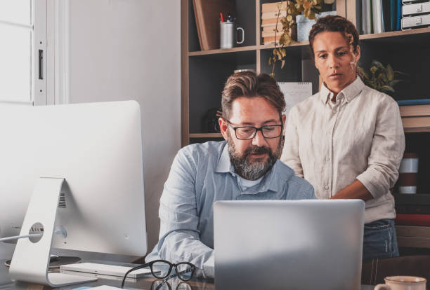 istock Mature caucasian businessman working with computer laptop in office with attractive curly woman standing learning 1370476604