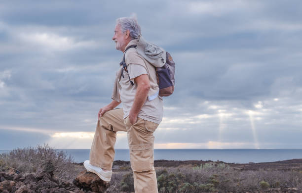 adulte caucasienne aux cheveux gris homme âgé faisant de la randonnée dans la campagne près de la mer portant un sac à dos regardant l’horizon en profitant de la nature. concept de liberté et de vacances. ciel nuageux. espace de copie - senior adult outdoors wellbeing sky photos et images de collection