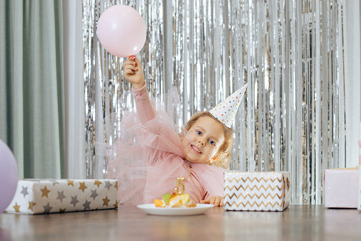 Cute little smiling girl with short curly hair and stars on face in pink dress and cap lift pink balloon near fruit cake with number five candle and gift boxes on tinsel background. Birthday concept.