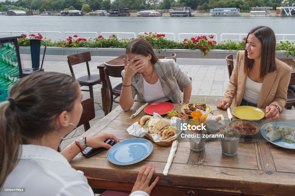Embarrassed young woman laughing with friends in outdoor restaurant serving Mexican snacks Young woman with hand over face looking embarrassed and laughing with friends, sitting in outdoor restaurant on city waterfront, Tex-Mex snacks on the table Embarrassment Stock Photo