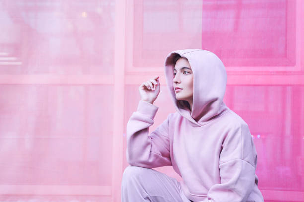 Młoda osoba ubrana w bluzę z kapturem siedzi w zamyśleniu na różowym tle.