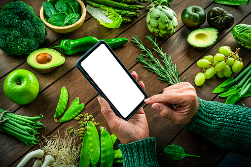 Close up of female hands holding blank screen mobile phone on a table full of green fruits and vegetables. High resolution 42Mp studio digital capture taken with SONY A7rII and Zeiss Batis 40mm F2.0 CF lens