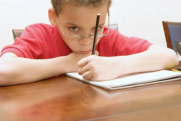Left handed young boy wearing glasses reluctant to do homework not paying attention, staring into space.