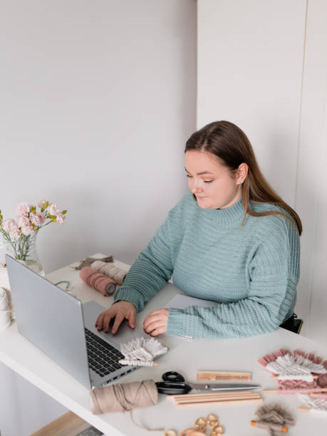 istock Young stylish woman using laptop at home, looking at screen, chatting, reading or writing email. Female hobby. Working on research project online concept. Handmade macrame shop 1370505991