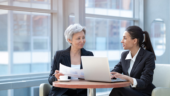 Two women analyzing documents while sitting on a table in office. Woman executives at work in office discussing some paperwork using laptop