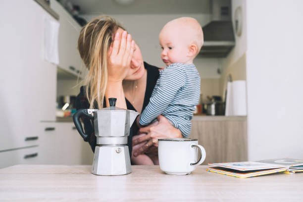 modern tired mother and little child after sleepless night. exhausted woman with baby is sitting with coffee in kitchen. life of working mother with baby. postpartum depression on maternity leave. - stereotypical housewife depression sadness women imagens e fotografias de stock