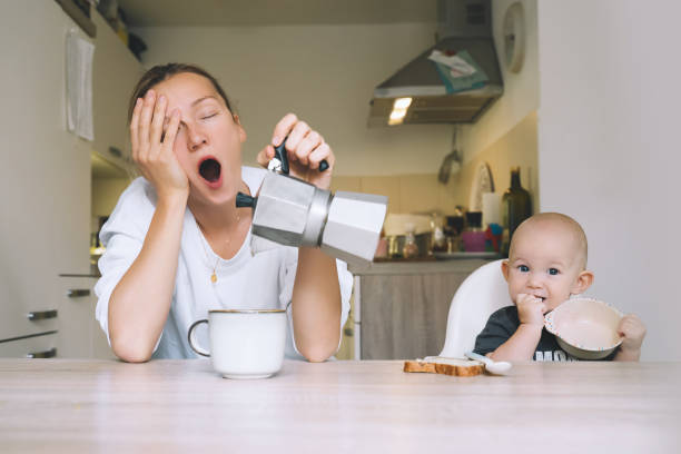 exhausted young woman with baby is sitting with coffee in kitchen. modern tired mom and little child after sleepless night. life of working mother with baby. postpartum depression on maternity leave. - stereotypical housewife depression sadness women imagens e fotografias de stock