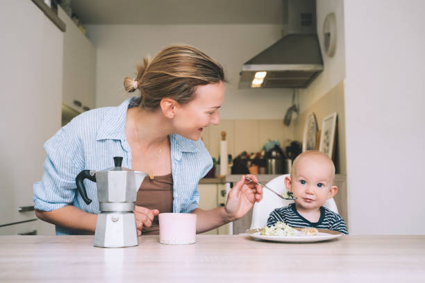 mãe sorridente e bebê adorando tomar café da manhã e se divertir na cozinha de casa. linda mãe aproveita sua vida de licença maternidade com seu filho. - mother green sparse contemporary - fotografias e filmes do acervo