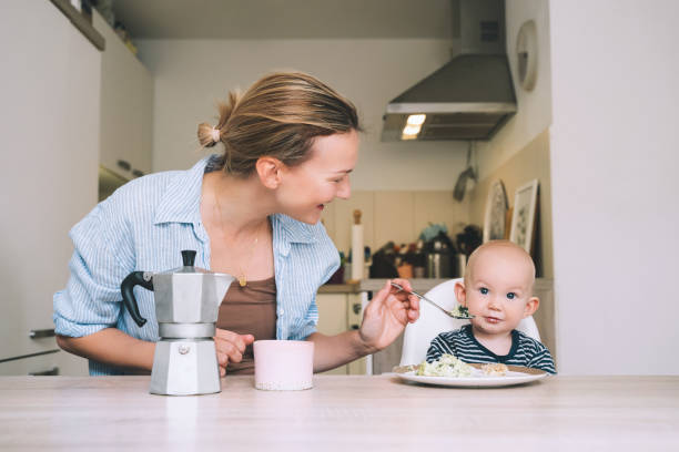 любящие улыбающиеся мама и малыш завтракают и веселятся на кухне дома. прекрасная мама наслаждается жизнью в декретном отпуске со своим ре� - mother green sparse contemporary стоковые фото и изображения