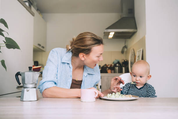 사랑하는 미소 짓는 어머니와 아기가 아침 식사를 하고 집에서 부엌에서 즐거운 시간을 보내고 있습니다. 아름다운 어머니는 자녀와 함께 출산 휴가에 그녀의 인생을 즐긴다. - mother green sparse contemporary 뉴스 사진 이미지