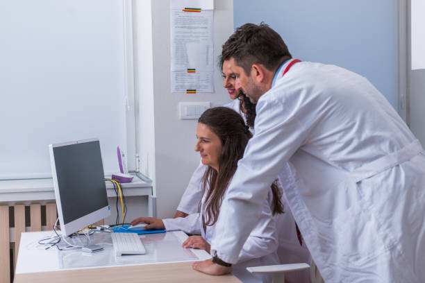 istock Two nurses ( female physicians ) and a male doctor standing next to a pc, mac ( desktop computer ) in the doctor's office while discussing a medical report. 1370509540