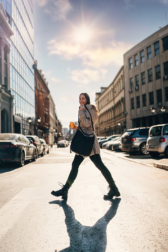 Happy young adult woman crossing city street on pedestrian crosswalk. She is smiled and looking over a shoulder while holding coffee to go in one hand and woman's bag in another. Bright sunny day.