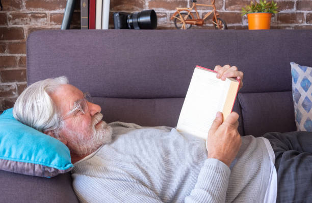 istock Senior attractive man lying down on sofa reading a book.  Old retired man relaxing at home.  Brick wall background 1370513863