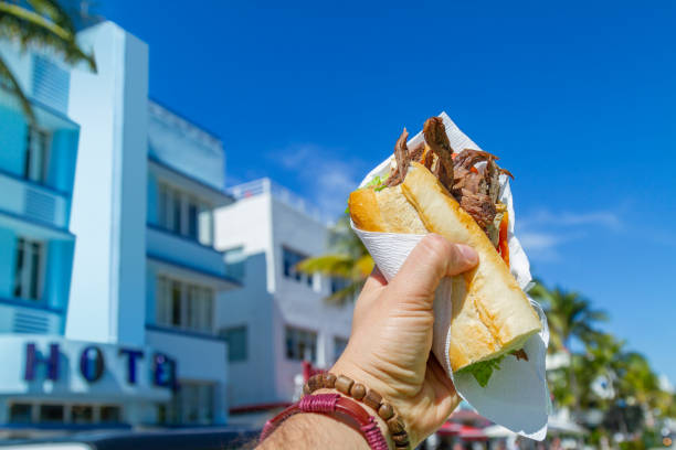 pov point of view aufnahme eines jungen reisemannes, der seine ferien genießt, während er ein kubanisches sandwich vor dem ocean drive, south beach, miami beach, miami, südflorida, vereinigte staaten von amerika isst. - kubaner stock-fotos und bilder