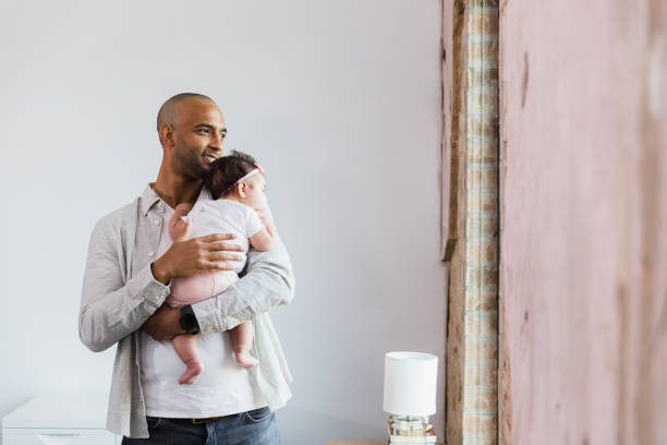 Happy new dad walks with baby in home The smiling mid adult father walks with his baby daughter in his home.  He looks out the living room window. 2 5 months stock pictures, royalty-free photos & images