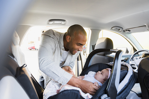 The new mid adult dad leans into the back of the car as he gently puts his baby daughter in her safety seat.