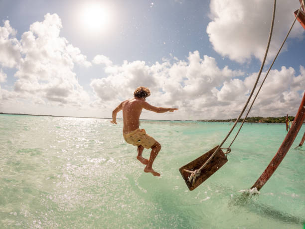 Young man jumps off a swing in a beautiful lagoon on a sunny day View of man swinging on white sand beach relaxing and sunbathing by the lagoon in Mexico swing play equipment stock pictures, royalty-free photos & images