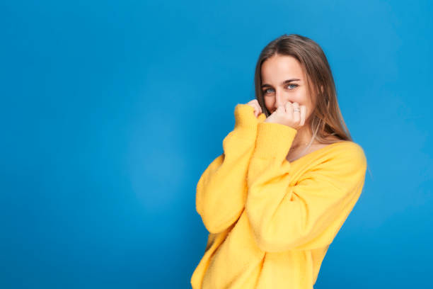 Young woman smiling shyly covers her face with hands. Isolated on blue background with yellow sweater. stock photo