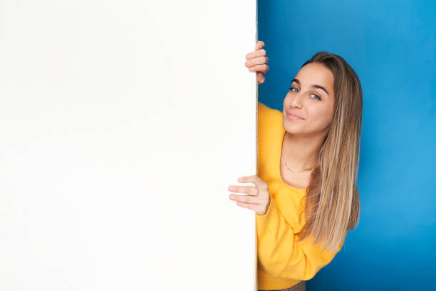 Portrait of a woman leaning out, hiding behind a white wall. isolated on a blue background. stock photo