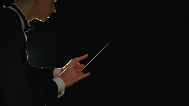 Symphony orchestra conductor wearing suit is directing musicians with movement of baton on black background with lights. Conducting, directing a musical performance with visible gestures. Close up