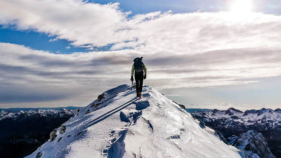 A man ski touring at  the top of the mountain.  He is showing his back an walking towards the peak. The view of the sky, clouds and mountains in the distance is seen in the background.