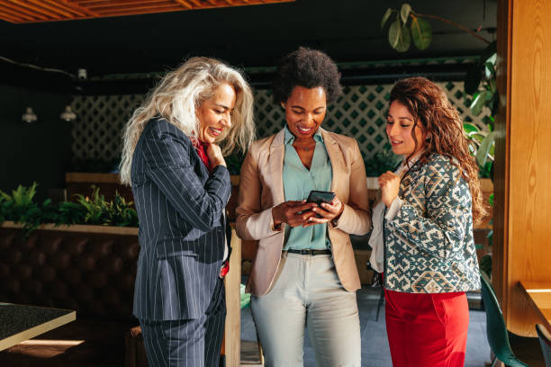 Three multiracial women using phone together Three happy beautiful multiracial women standing at cafe chatting talking playing with cellphone in coffee shop or tea house. international match stock pictures, royalty-free photos & images