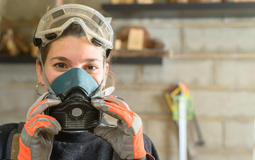 Woman in protective transparent googles and respirator looking at camera while standing in light workshop with professional equipment during woodwork
