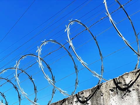 A barbed wire with an electric fence on a wall, resembling a prison wall or a border wall to prevent escapes or trespassing, with a blue sky in the background
