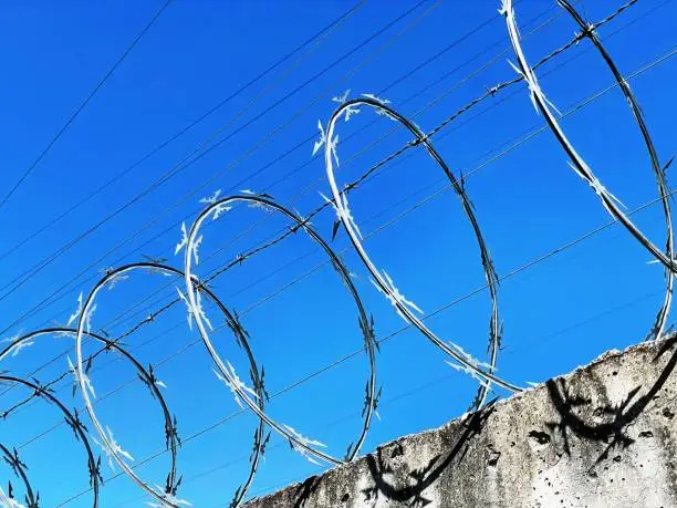 A barbed wire with an electric fence on a wall, resembling a prison wall or a border wall to prevent escapes or trespassing, with a blue sky in the background