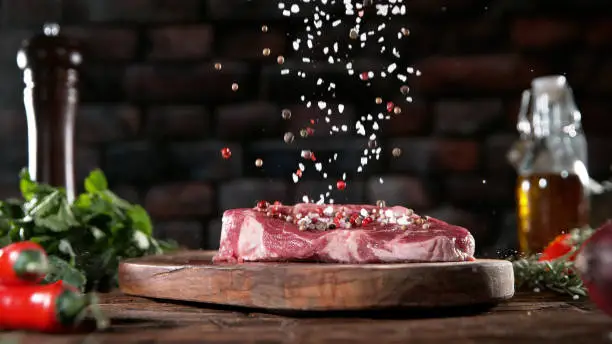 Falling grained salt and pepper spice on raw meat steak, placed on wooden cutting board. Preparation of meat, dark background.