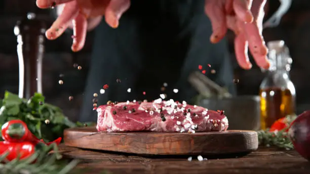 Chef pouring grained salt and pepper spice on raw meat steak, placed on wooden cutting board. Preparation of meat, dark background.