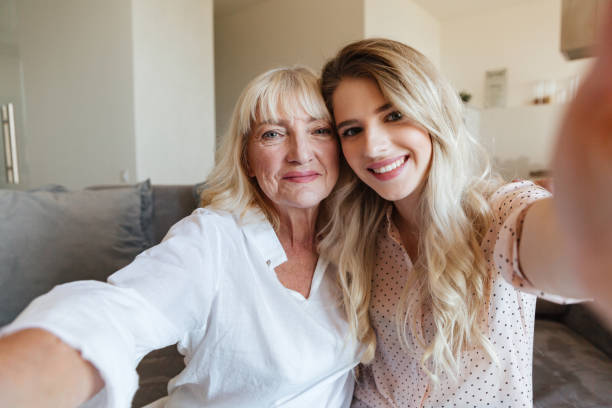 istock Cheerful lady on sofa with grandmother hugging make selfie. 1370549323