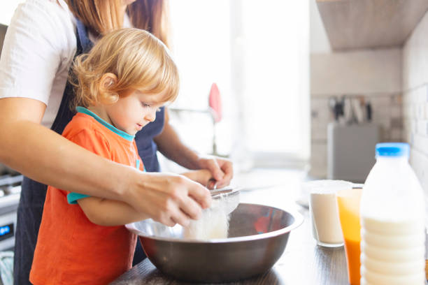 family in a kitchen. mother and child sieving flour into a bowl. mom and small kid son cooking together making dough in bowl in kitchen. . homemade food and a little helper - chef cookie dishware domestic kitchen imagens e fotografias de stock