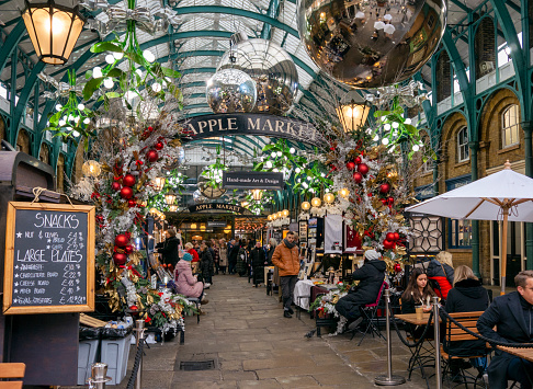 Tourists, shoppers and diners in the former Apple Market inside London’s popular Covent Garden Market before Christmas. The market is bright with festive decorations.